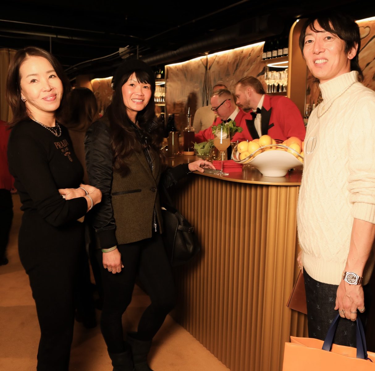 Three people smiling at the camera with bartenders preparing drinks in the background Three people smiling at the camera with bartenders preparing drinks in the background